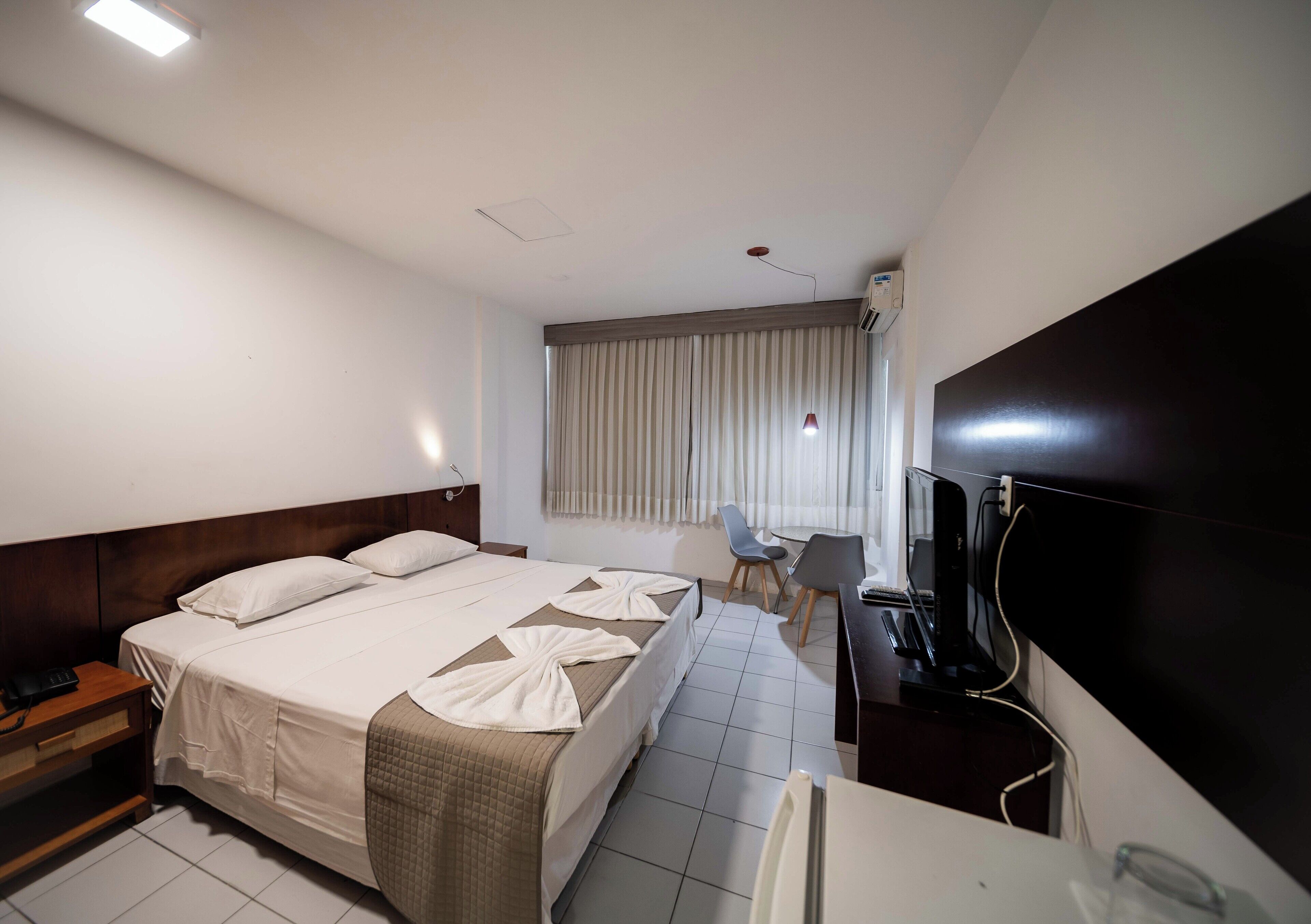 A hotel room with a bed, bedside table, desk, chair, TV, and a window with curtains.