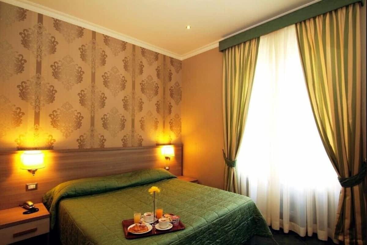Ein Hotelzimmer mit einem großen Bett, zwei Nachttischlampen, einem Tablett mit Frühstücksgegenständen und einem Fenster mit Vorhängen.