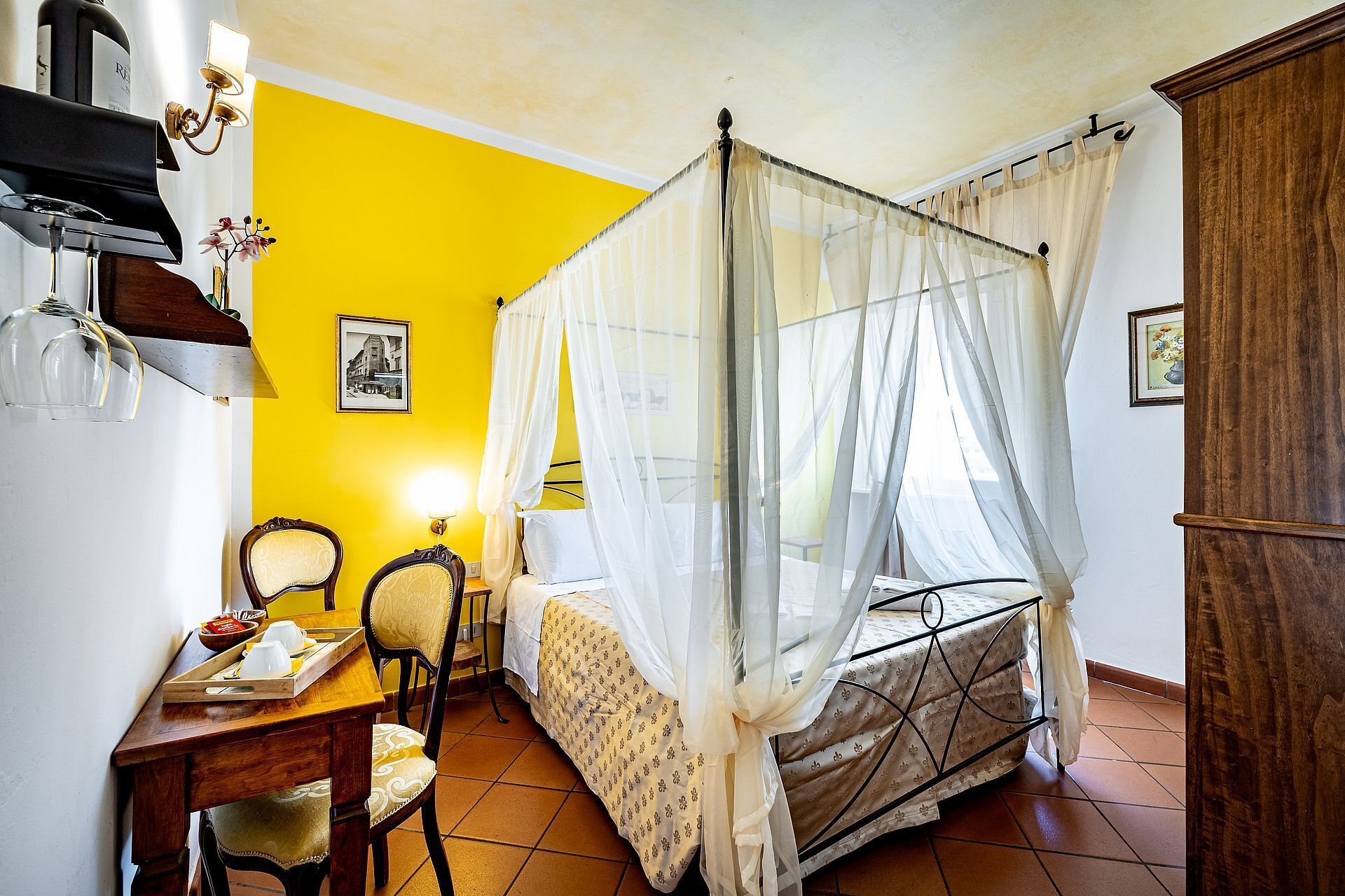 A room with a bed, a desk with chairs, a lamp, and a picture on the wall.