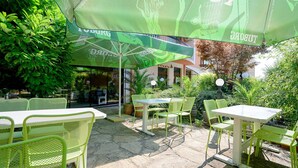 An outdoor patio area with green chairs and tables, covered by large green umbrellas, and surrounded by lush greenery. - Royal Beach (Varna)