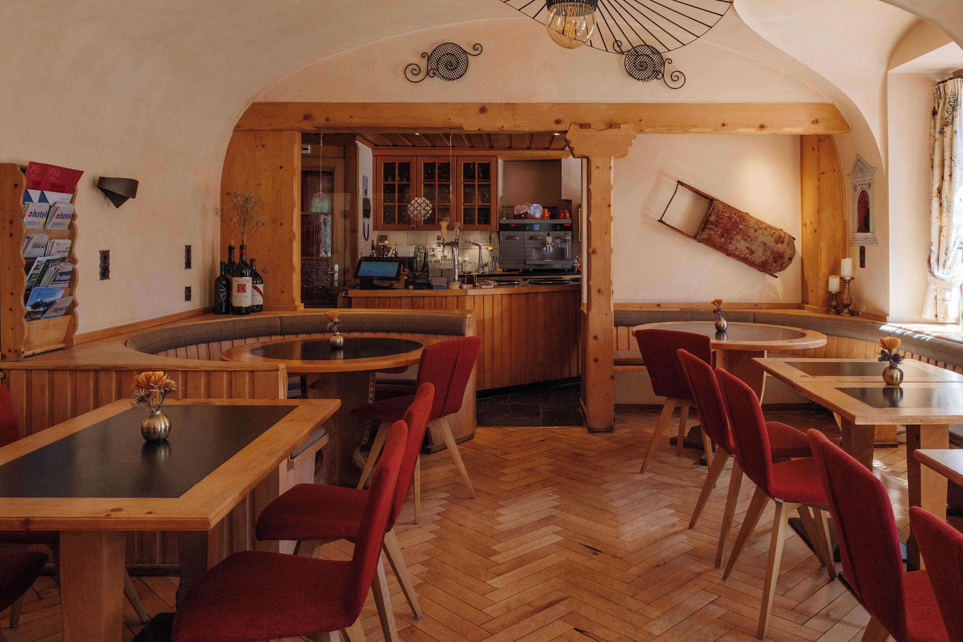 A restaurant with wooden tables and red chairs, a kitchen in the background, and a herringbone floor pattern.