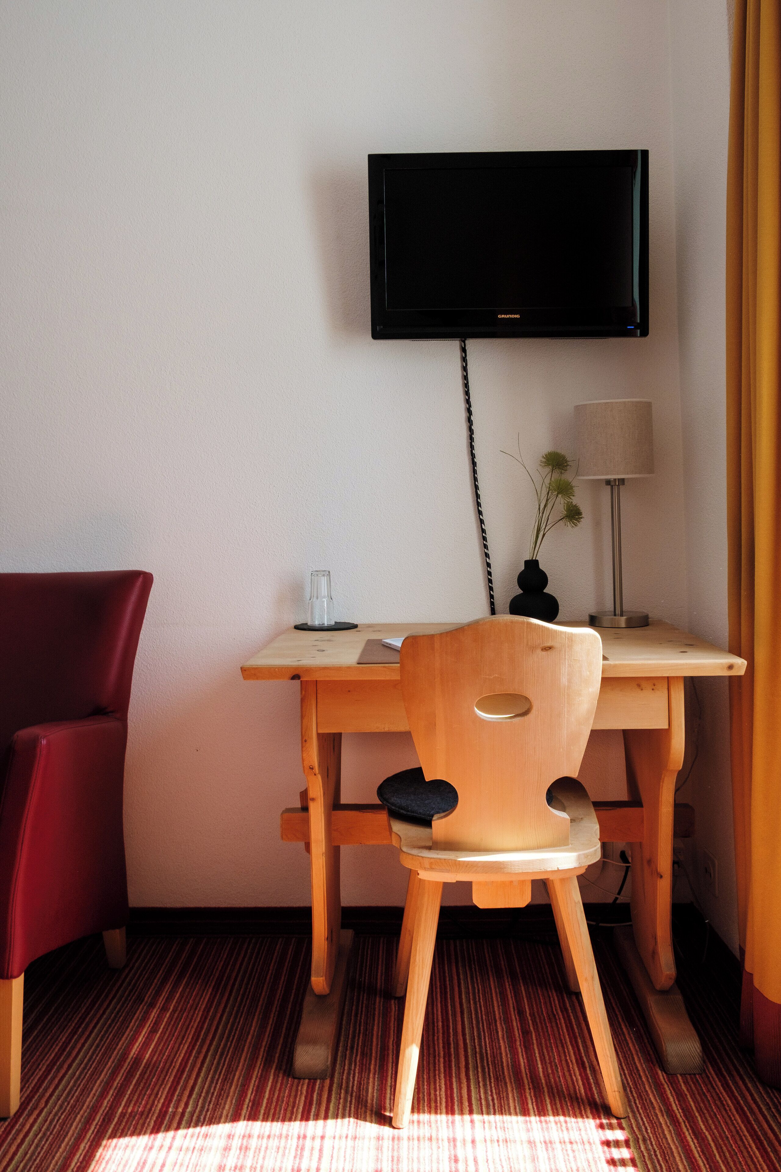 A room with a wooden desk, a red chair, a television mounted on the wall, and a lamp on the desk.