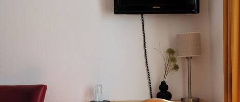 A room with a wooden desk, a red chair, a television mounted on the wall, and a lamp on the desk.