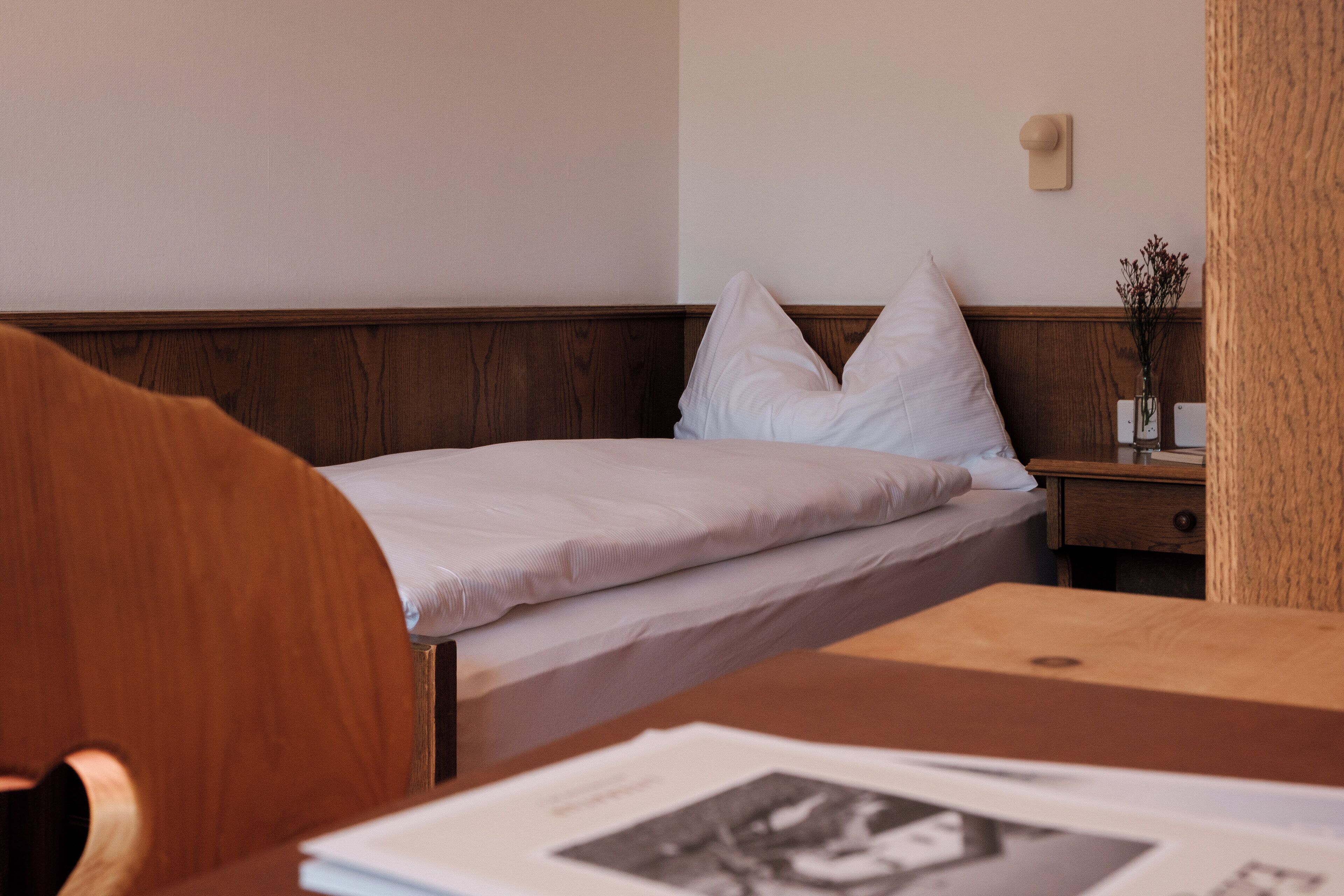 Een hotelkamer met een houten hoofdeinde, een netjes opgemaakt bed met witte lakens, een nachtkastje met een bloemenvaas en een klein nachtkastje.