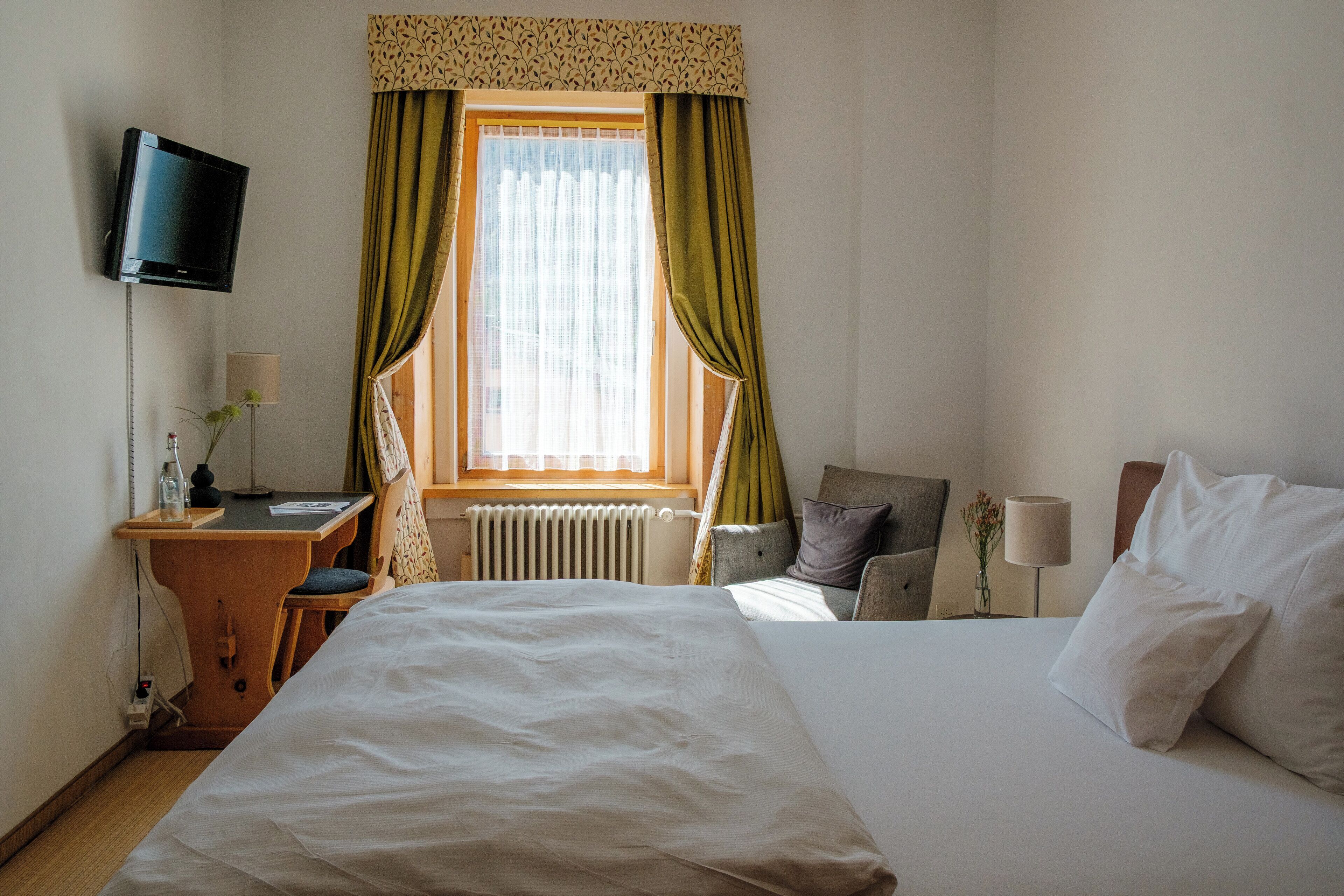 A hotel room with a bed, a desk, a chair, a TV, and a window with curtains.