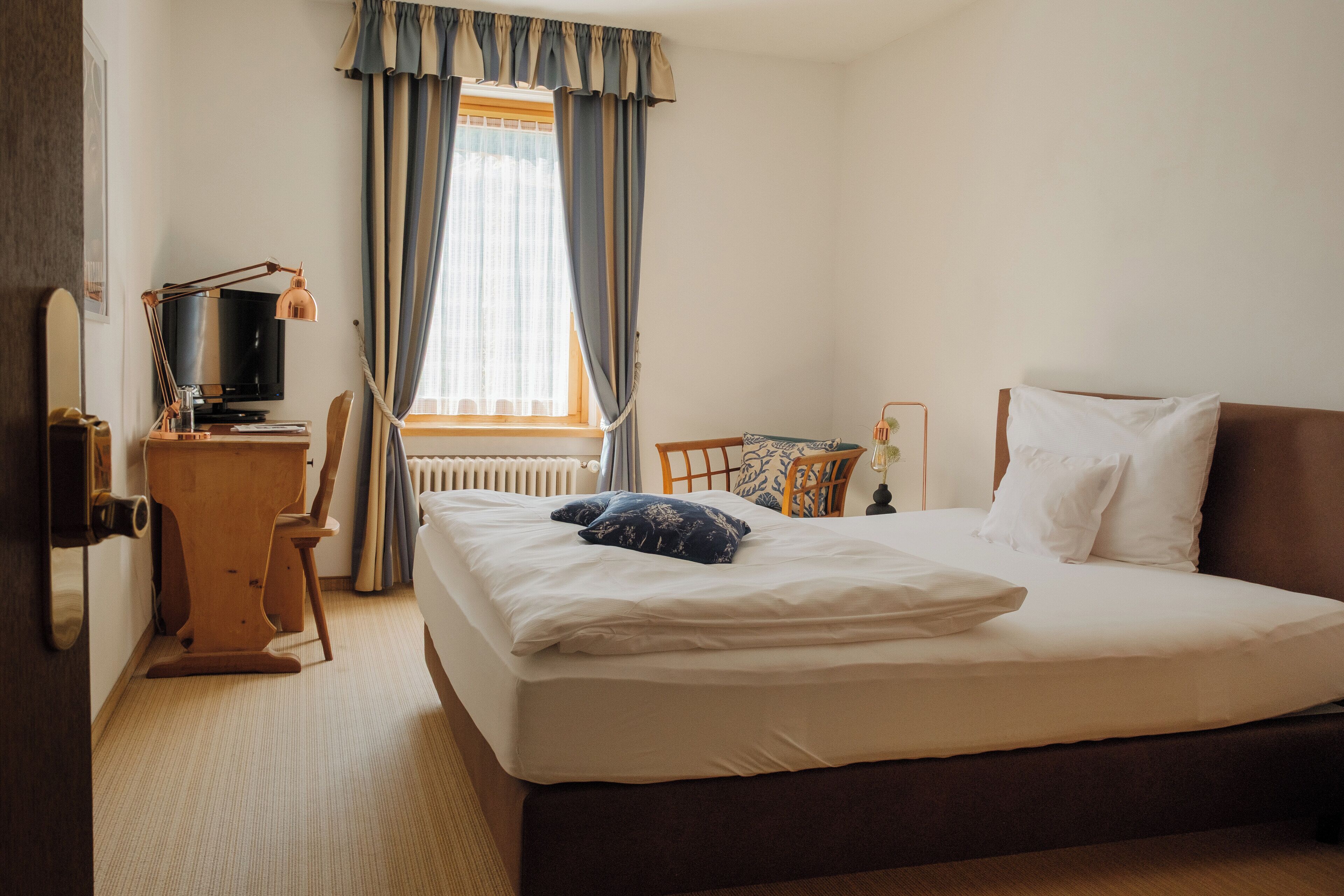 A hotel room with a large bed, a desk with a computer, a chair, and a window with curtains.