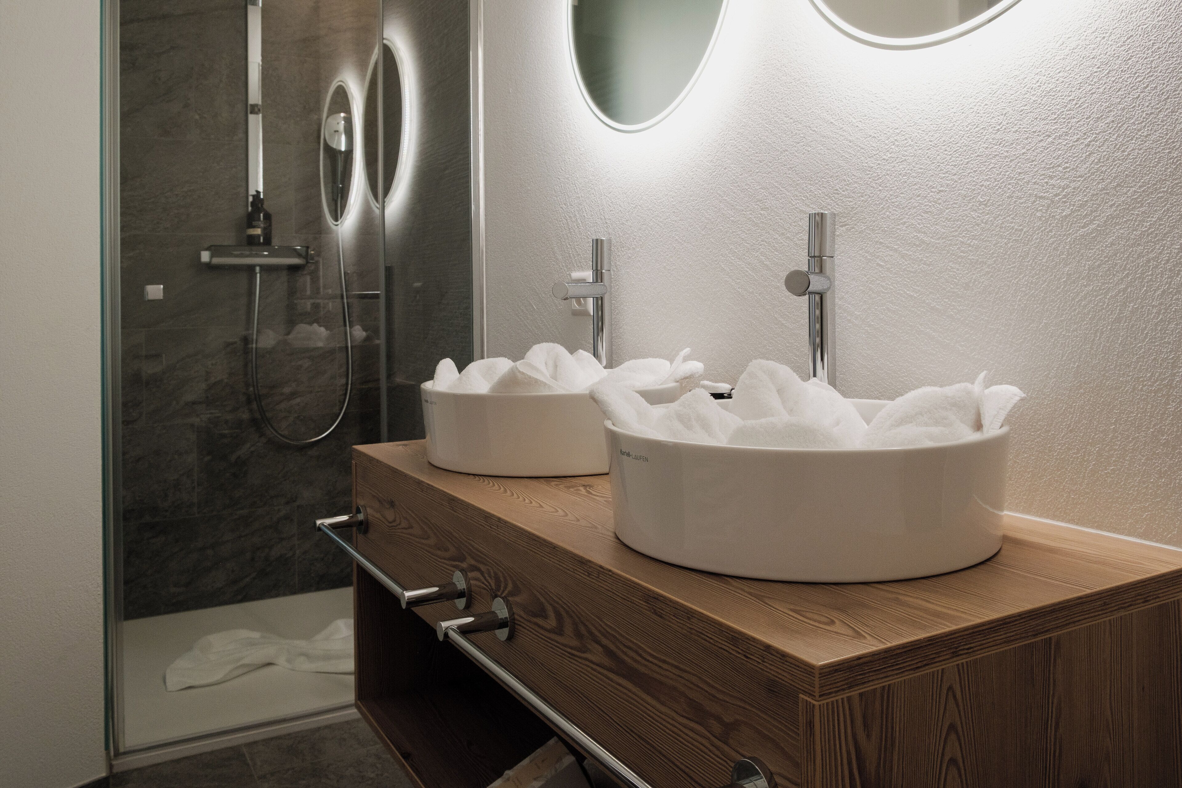 A modern bathroom with two round white sinks, a wooden vanity, and a glass shower enclosure.