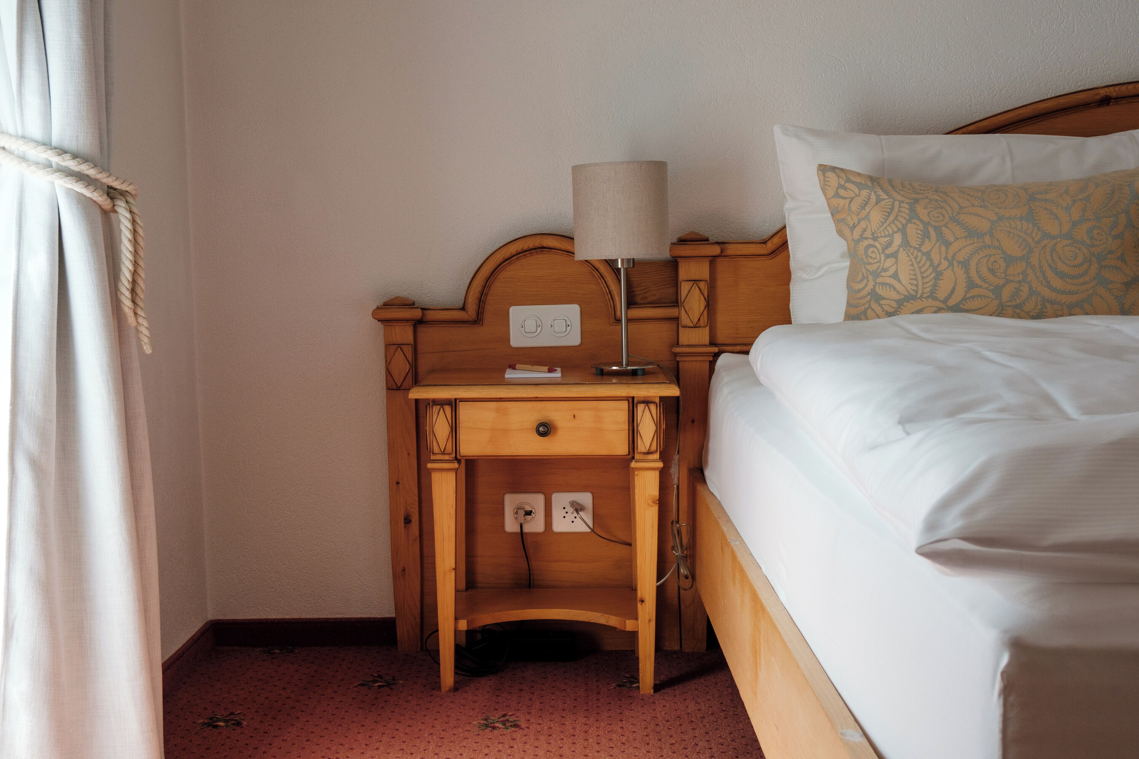 A neatly made bed with a wooden nightstand beside it, featuring a lamp and a power outlet.