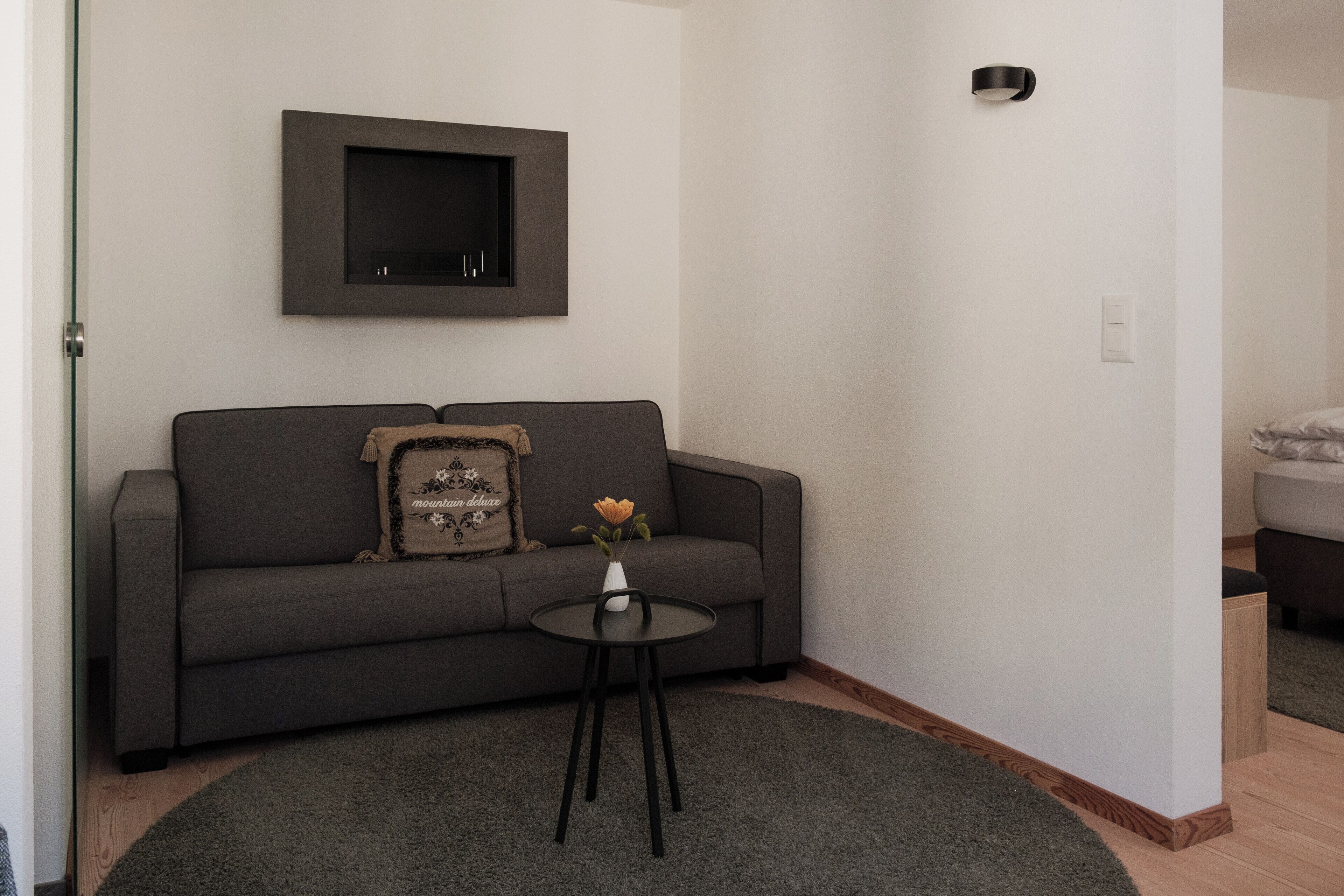 A modern living room with a grey sofa, a small black table with a vase of flowers, and a mounted TV on the wall.
