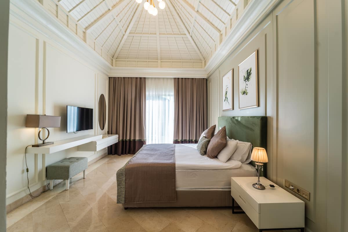 A modern hotel room with a large bed, a TV, a bedside table with a lamp, and a bench.