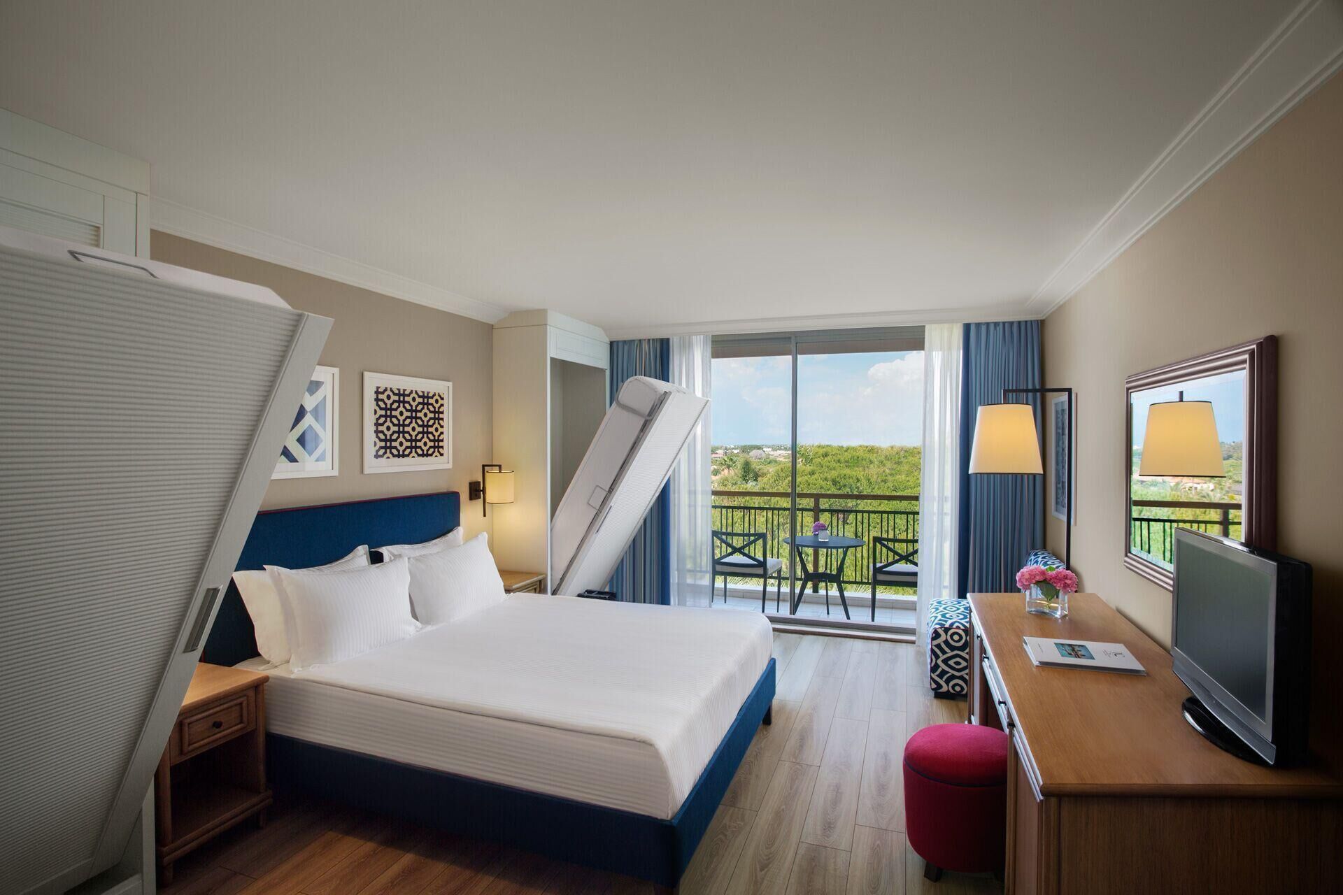 Habitación de hotel con cama, escritorio con televisor, balcón con mesa y sillas, y una escalera.