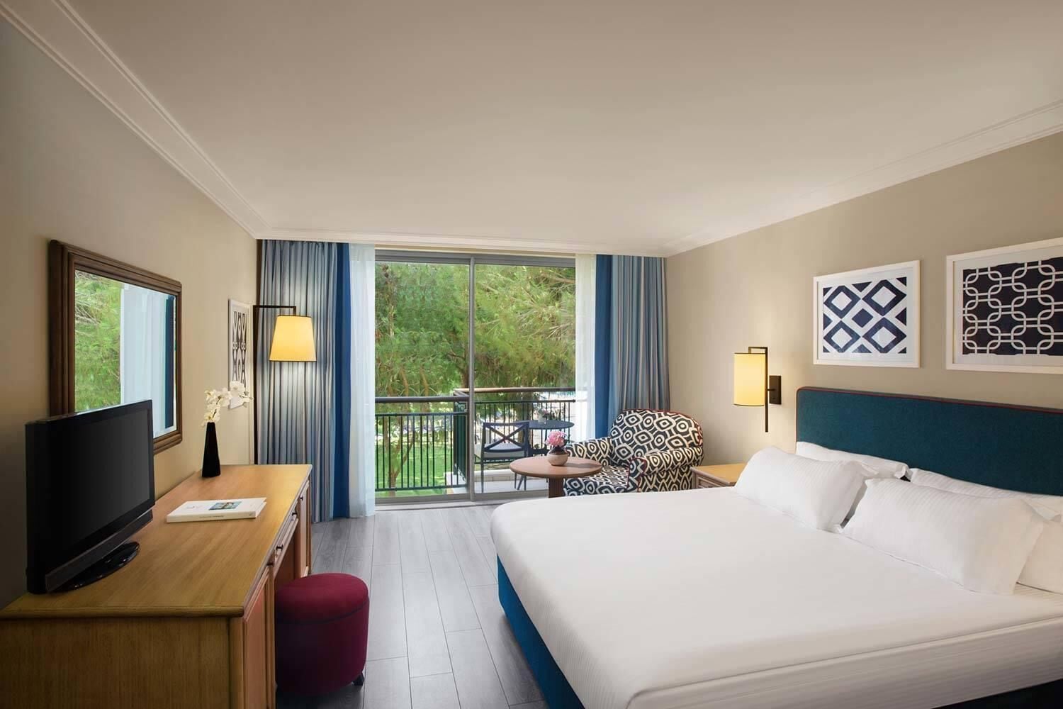 A hotel room with a bed, a desk with a TV, a chair, a balcony with a view of trees, and two wall-mounted lamps.
