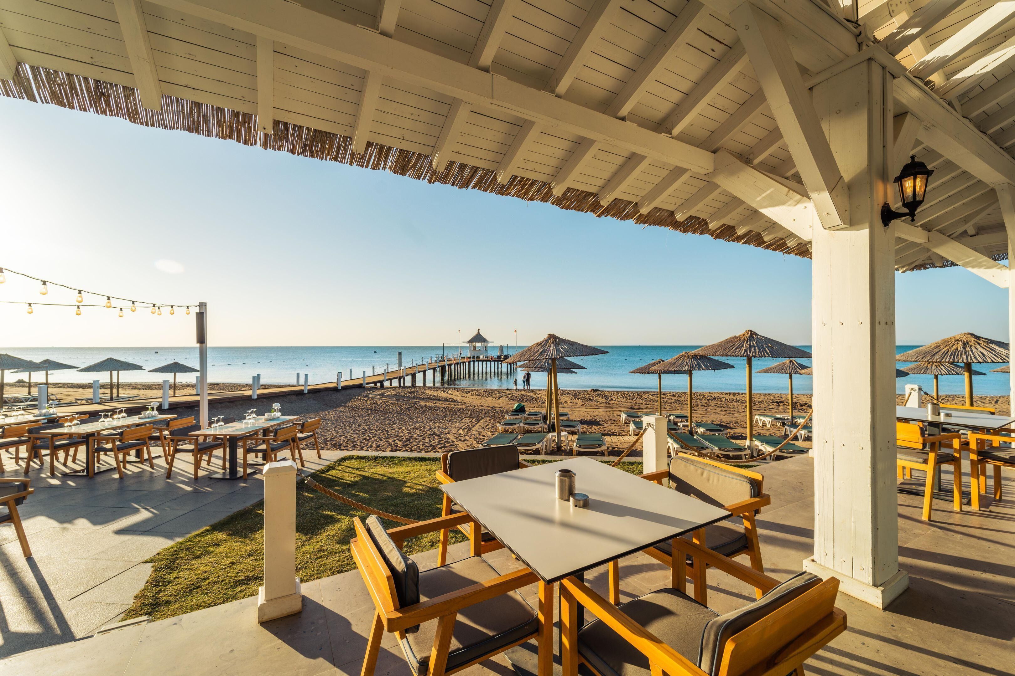 Een terras met tafels en stoelen met uitzicht op een strand en een pier.