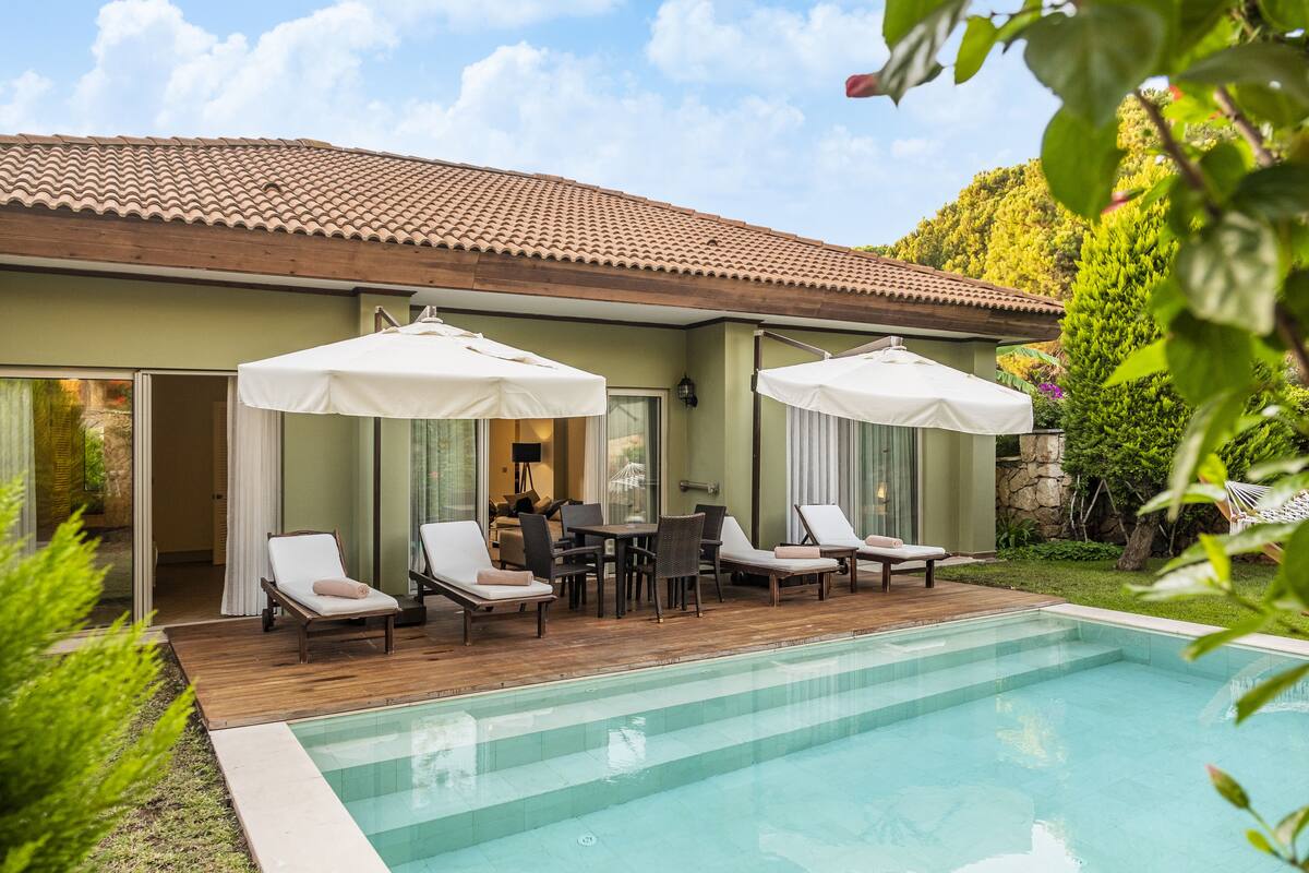 A poolside area with lounge chairs and umbrellas, a wooden deck, and a building with a tiled roof.