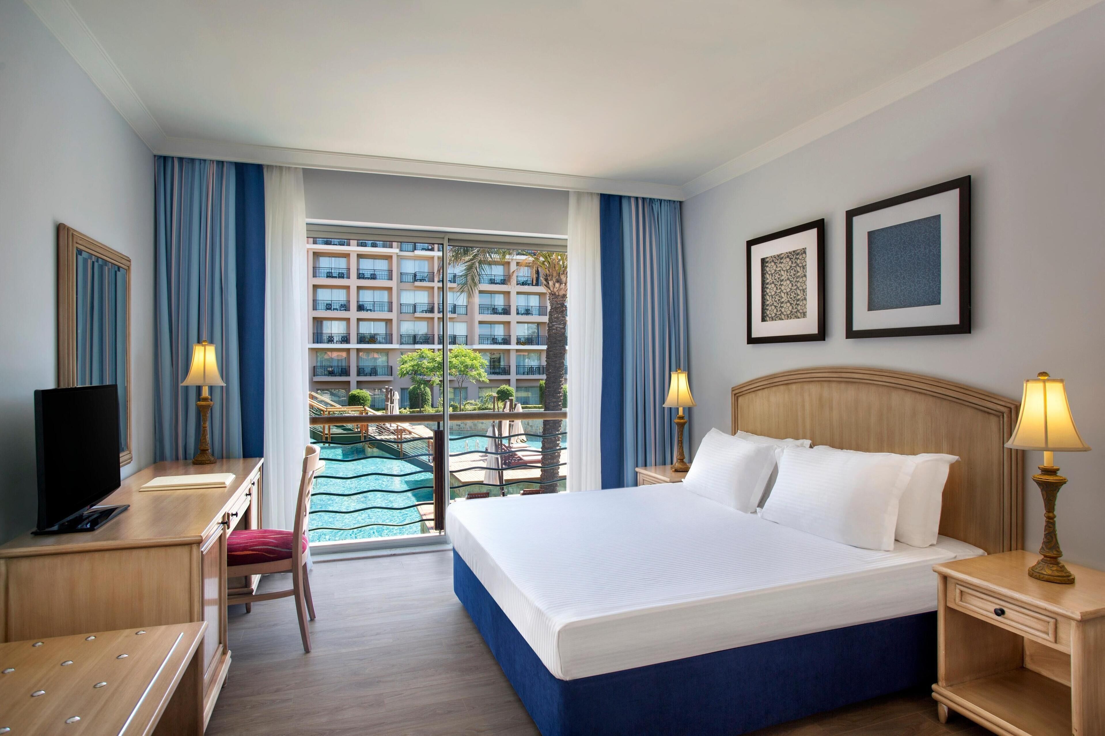 Une chambre d’hôtel avec un lit, un bureau, une télévision et une vue sur une piscine et des immeubles.