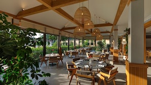 A spacious dining area with wooden beams and large windows overlooking a garden. Tables are set with chairs, and there are hanging wicker lamps. - IC Hotels Green Palace & Villas - All inclusive (Antalya)