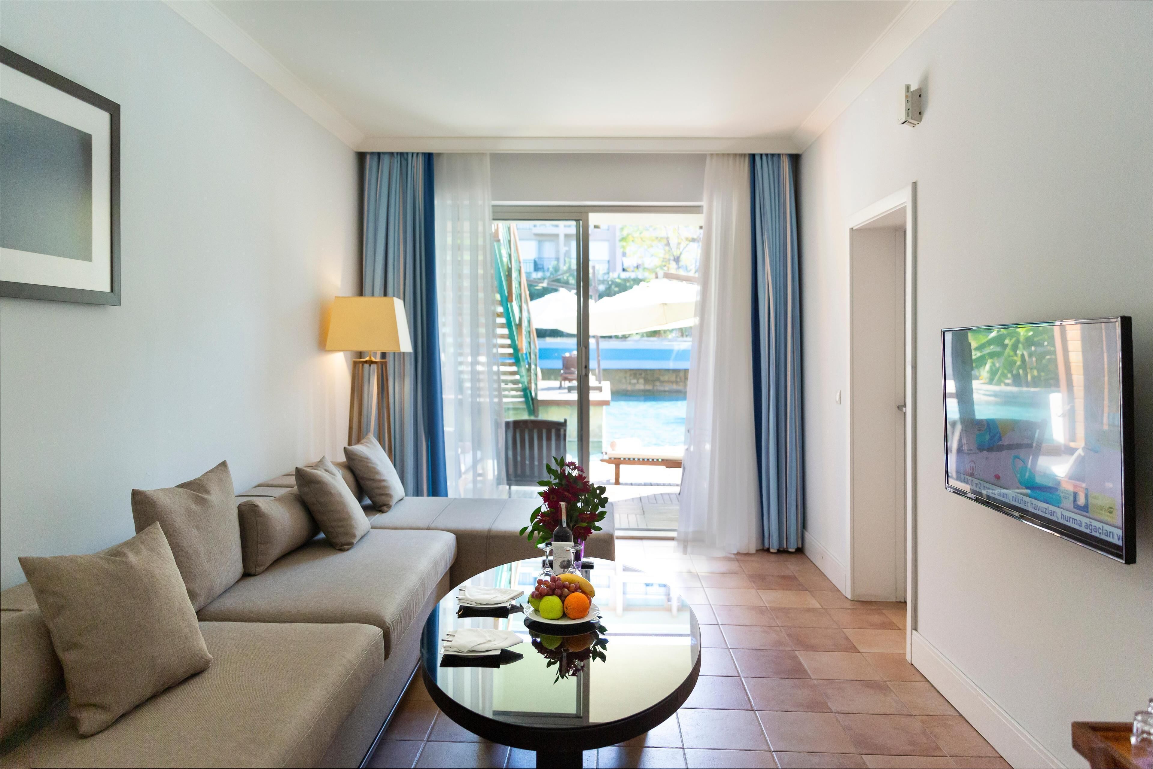 Una camera d'albergo moderna con un divano, un tavolino in vetro con della frutta, una TV e vista su una piscina attraverso una porta in vetro.