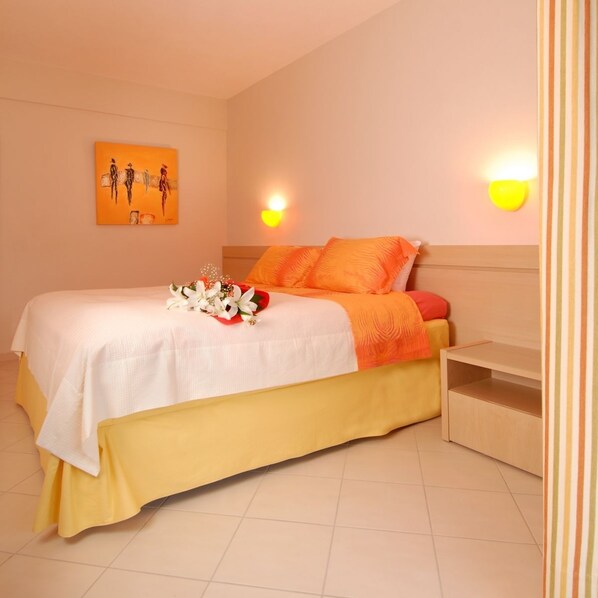 Una cama bien hecha con una colcha blanca y naranja, un ramo de flores y un cabecero con dos luces fijadas a la pared.