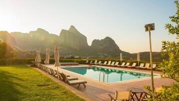 A pool area with lounge chairs and umbrellas, surrounded by greenery and mountains in the background.