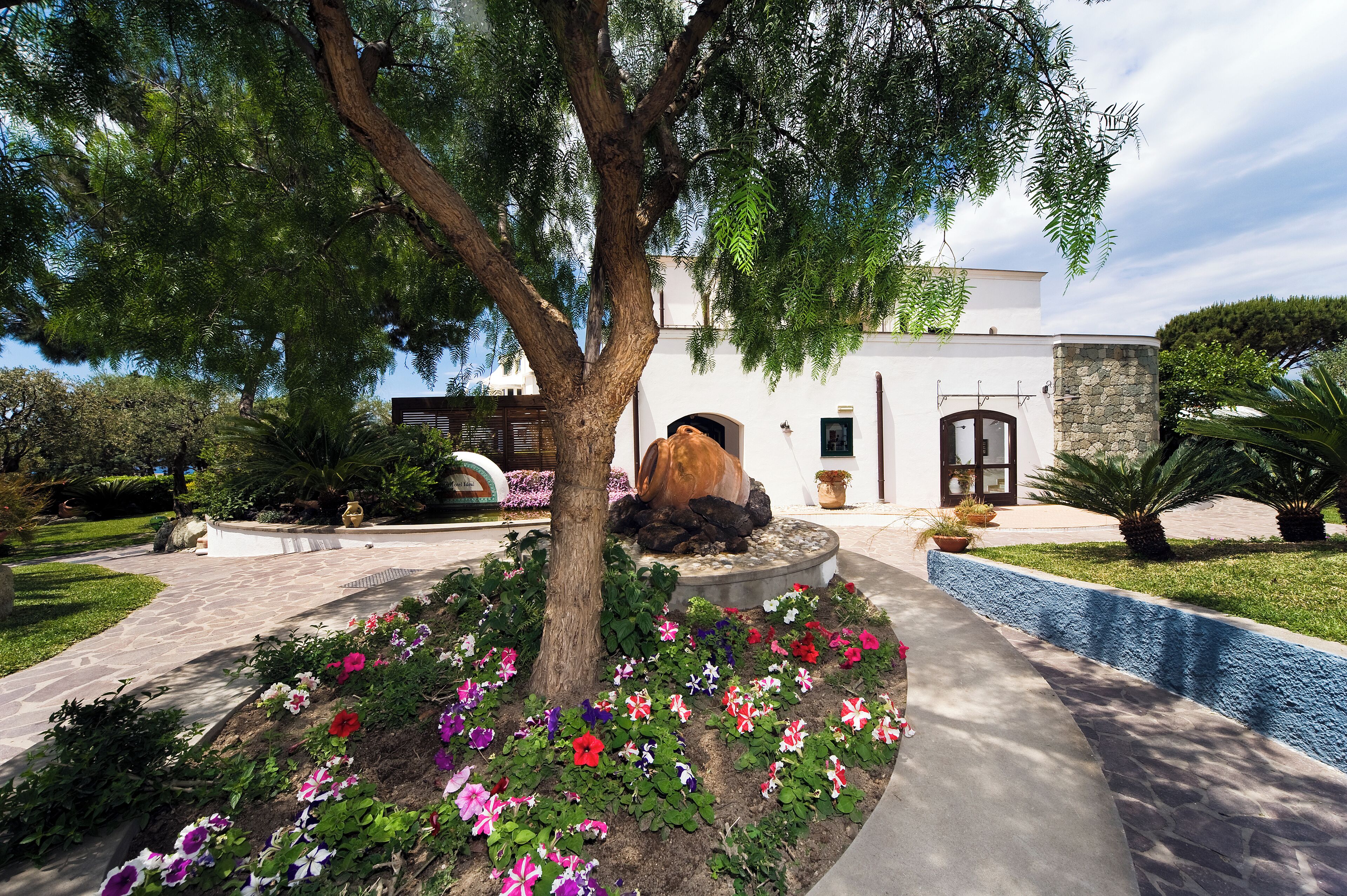 A modern building with a stone facade, surrounded by a landscaped garden with colorful flowers and a large tree.
