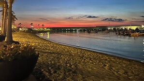 Une plage au crépuscule, avec une mer calme, des palmiers et une jetée en arrière-plan.