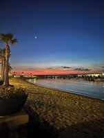 Una playa al atardecer con mar tranquilo, palmeras y un muelle al fondo.