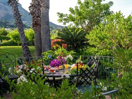 Um jardim com mesa e cadeiras, cercado por vegetação exuberante e montanhas ao fundo.