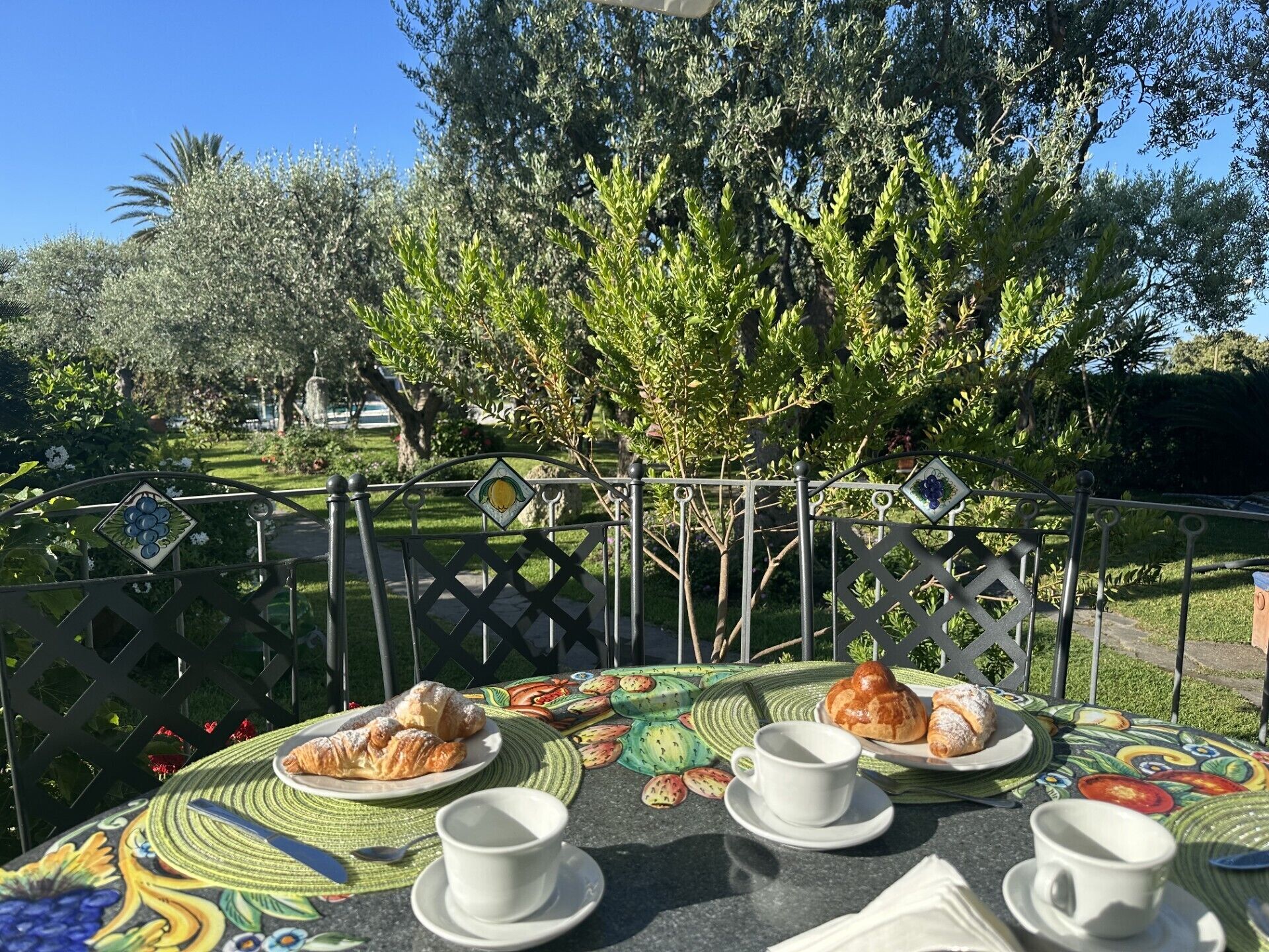 Une table placée à l’extérieur, avec des croissants et des tasses de café, entourée d’un jardin arboré et d’une clôture en métal.