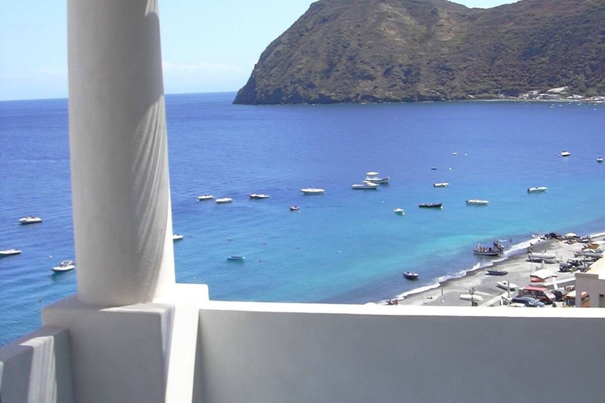 A balcony with a view of the sea and boats, a white pillar, and a tiled floor.