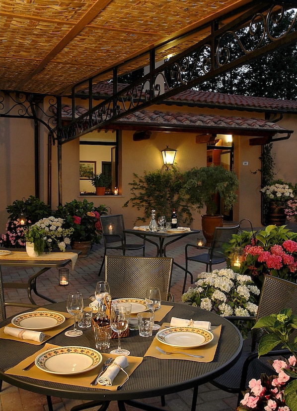 An outdoor dining area with round tables set for two, featuring metal chairs and a pergola overhead.