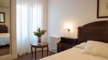 A hotel room with a bed, a bedside table with a vase of flowers, a radiator, and a wall-mounted light fixture.