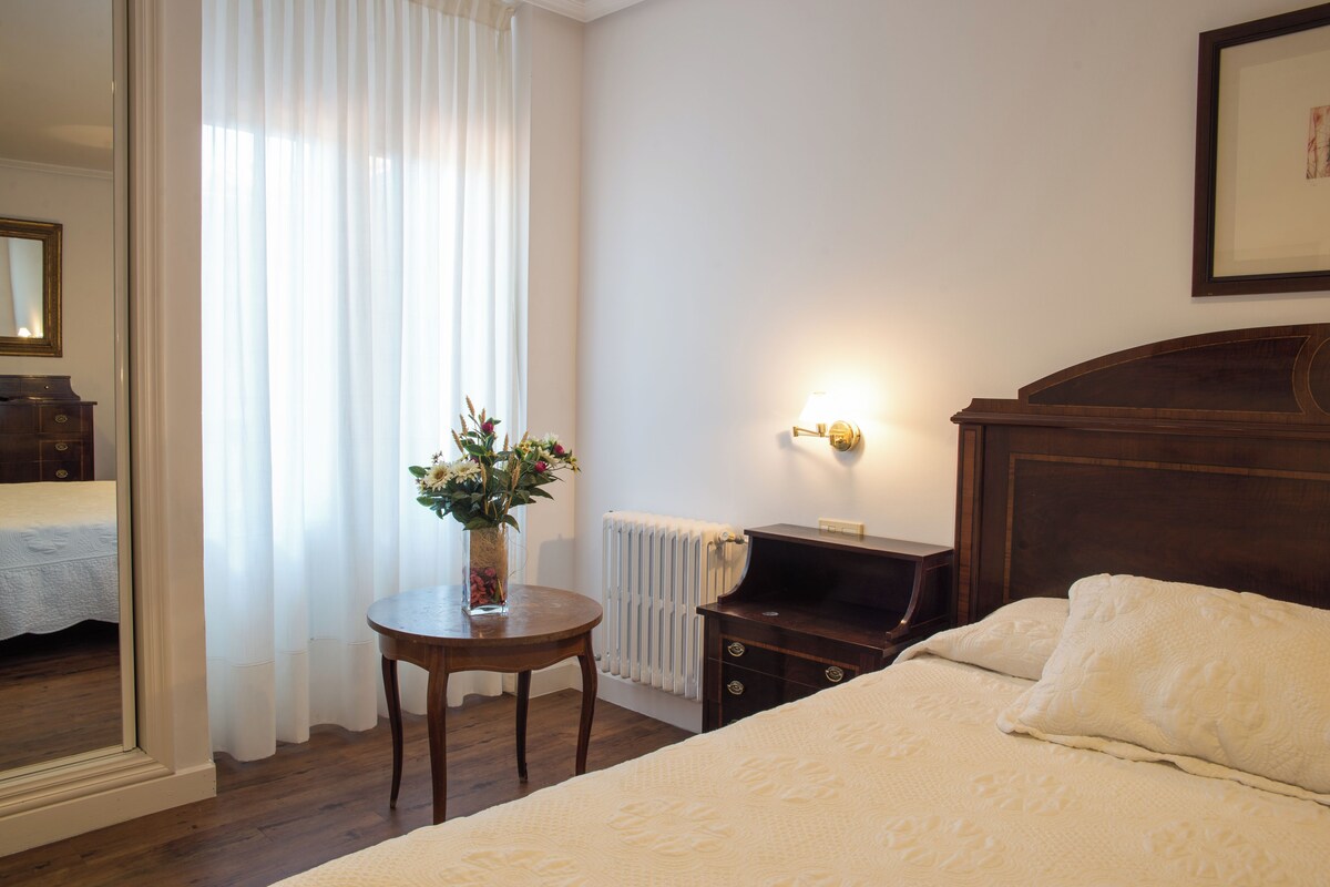 Ein Schlafzimmer mit hölzernem Kopfteil, einem Nachttisch mit Blumenvase, einem Heizkörper und einem gerahmten Bild an der Wand.