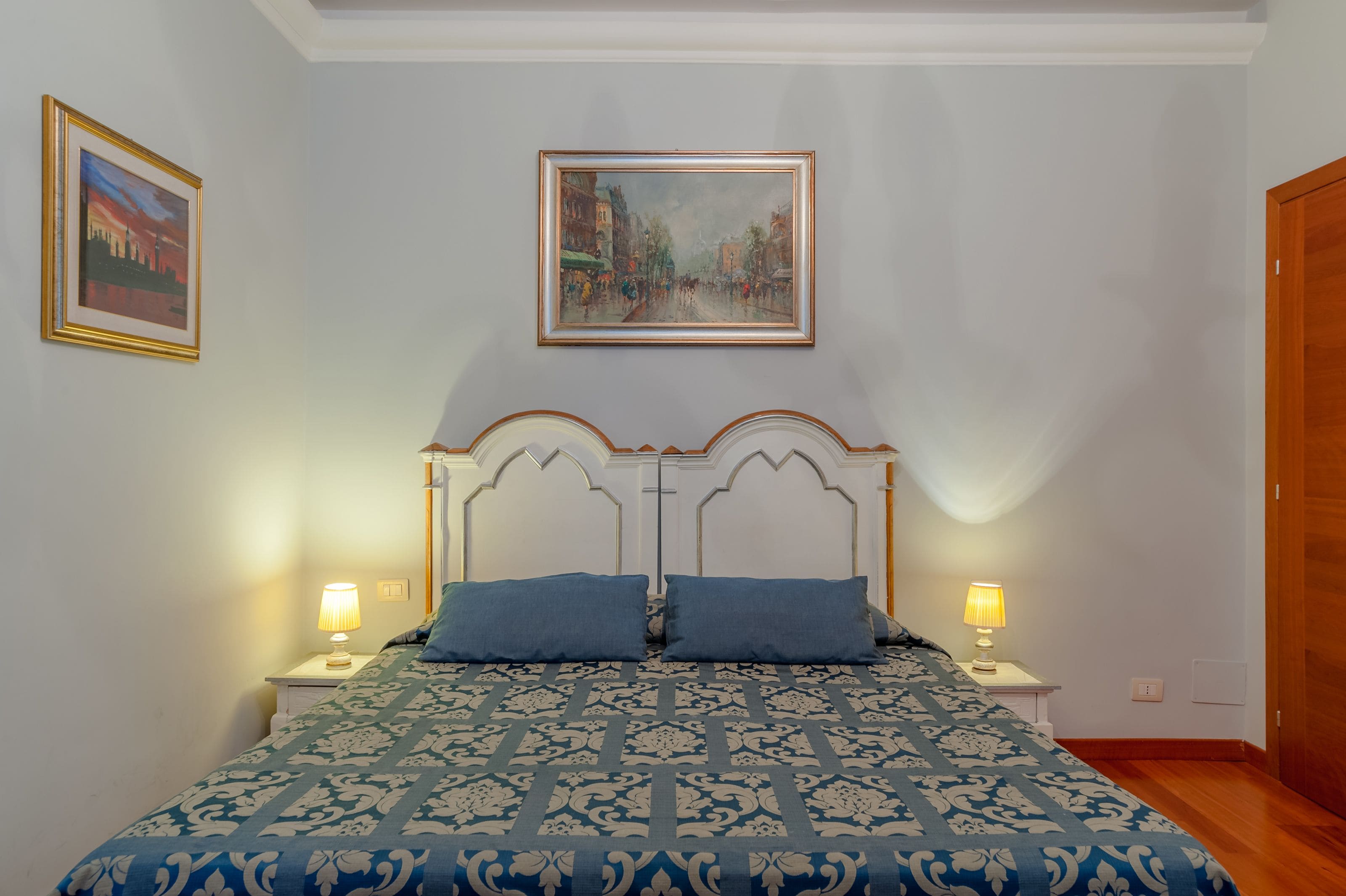 A bedroom with a bed, two bedside lamps, a framed painting above the bed, and a wooden door.