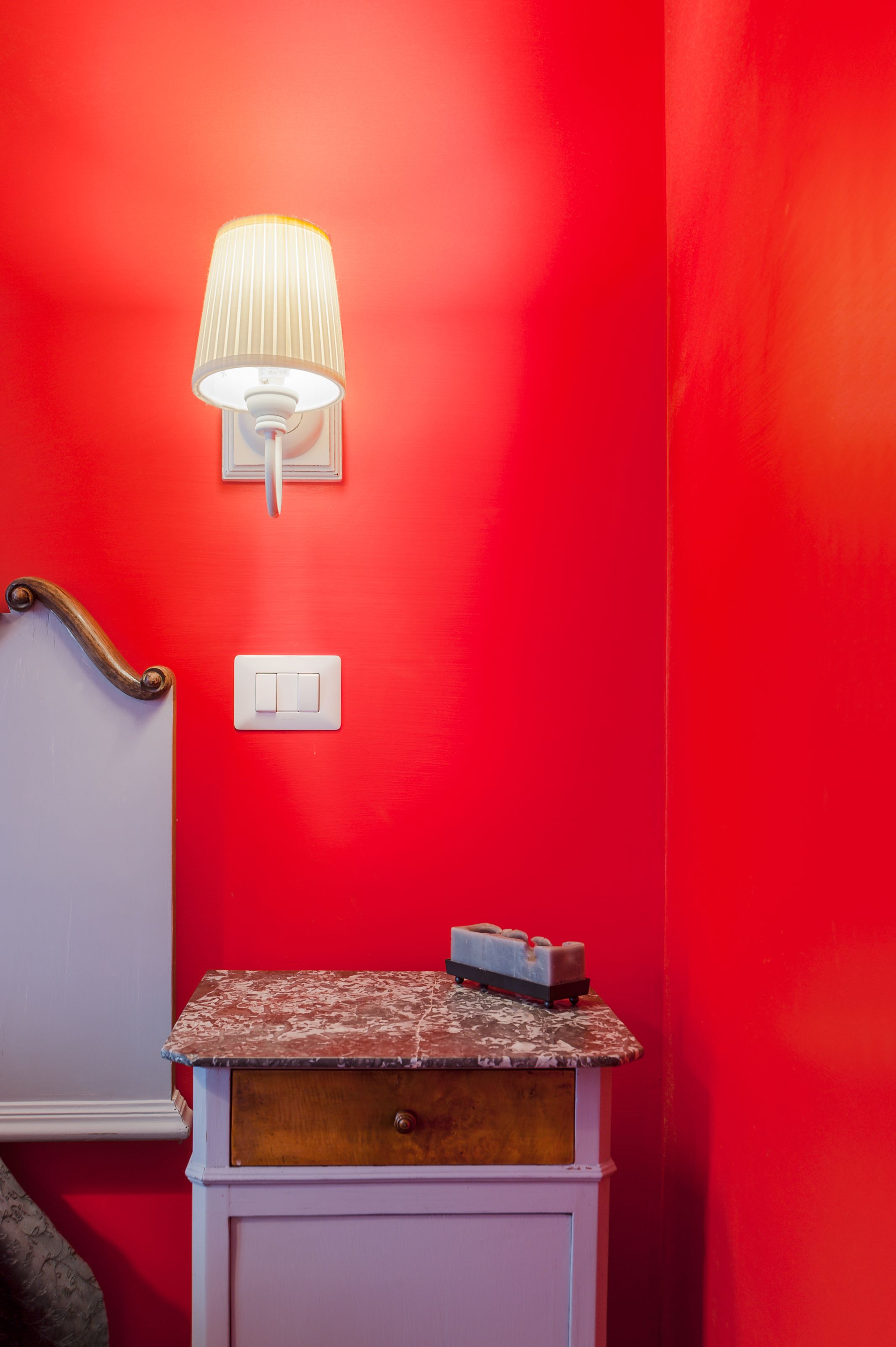 Eine rote Wand mit einem weißen Lichtschalter, einer Wandleuchte und einem kleinen Nachttisch mit Schublade.