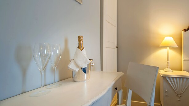 Ein Zimmer mit einem weißen Tisch, auf dem zwei Weingläser und eine Champagnerflasche stehen. Auf einem Nachttisch steht eine Lampe und neben dem Tisch ein Sessel.