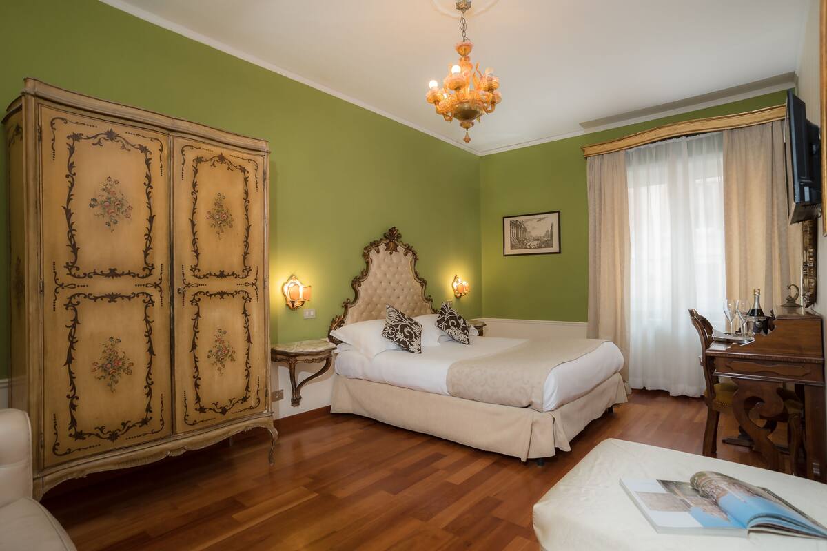 Ein Schlafzimmer mit großem Holzschrank, Bett mit weißer Bettwäsche, einer Chandelierleuchtre und einem Schreibtisch mit Stuhl.