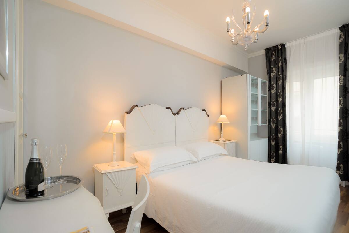 Ein Hotelzimmer mit einem großen Bett, zwei Nachttischen mit Lampen, einer Ablage mit Flasche und Gläsern, einem Spiegel und einer Chandelier.