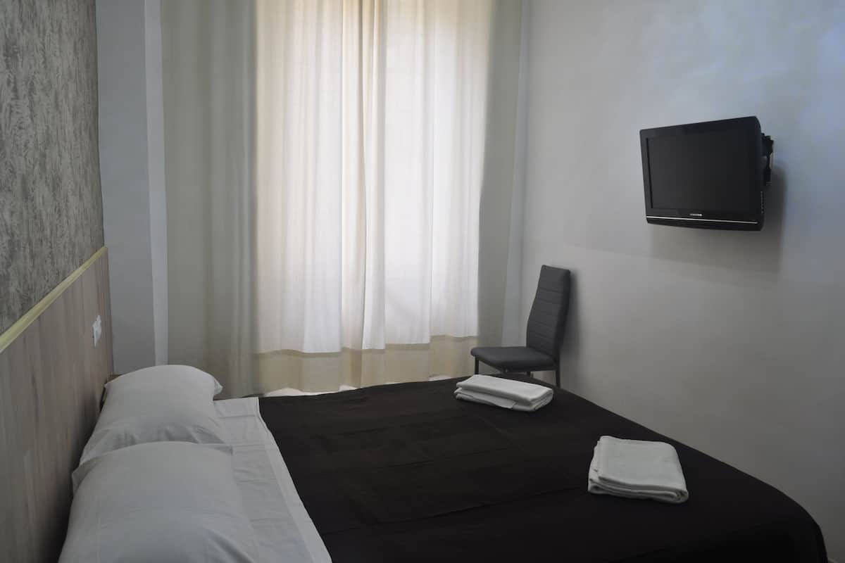 A hotel room with a bed, a chair, a TV mounted on the wall, and a window with curtains.