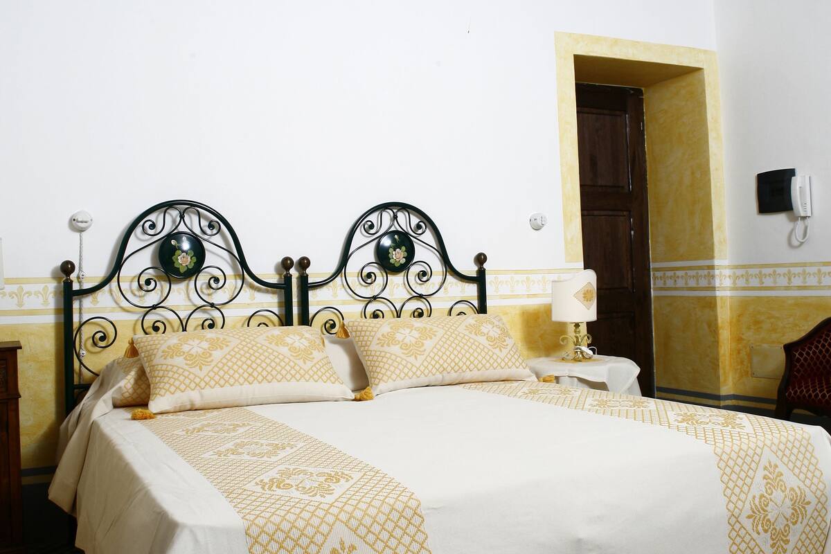 A bed with a patterned bedspread, a bedside table with a lamp, and a wooden door in the background.