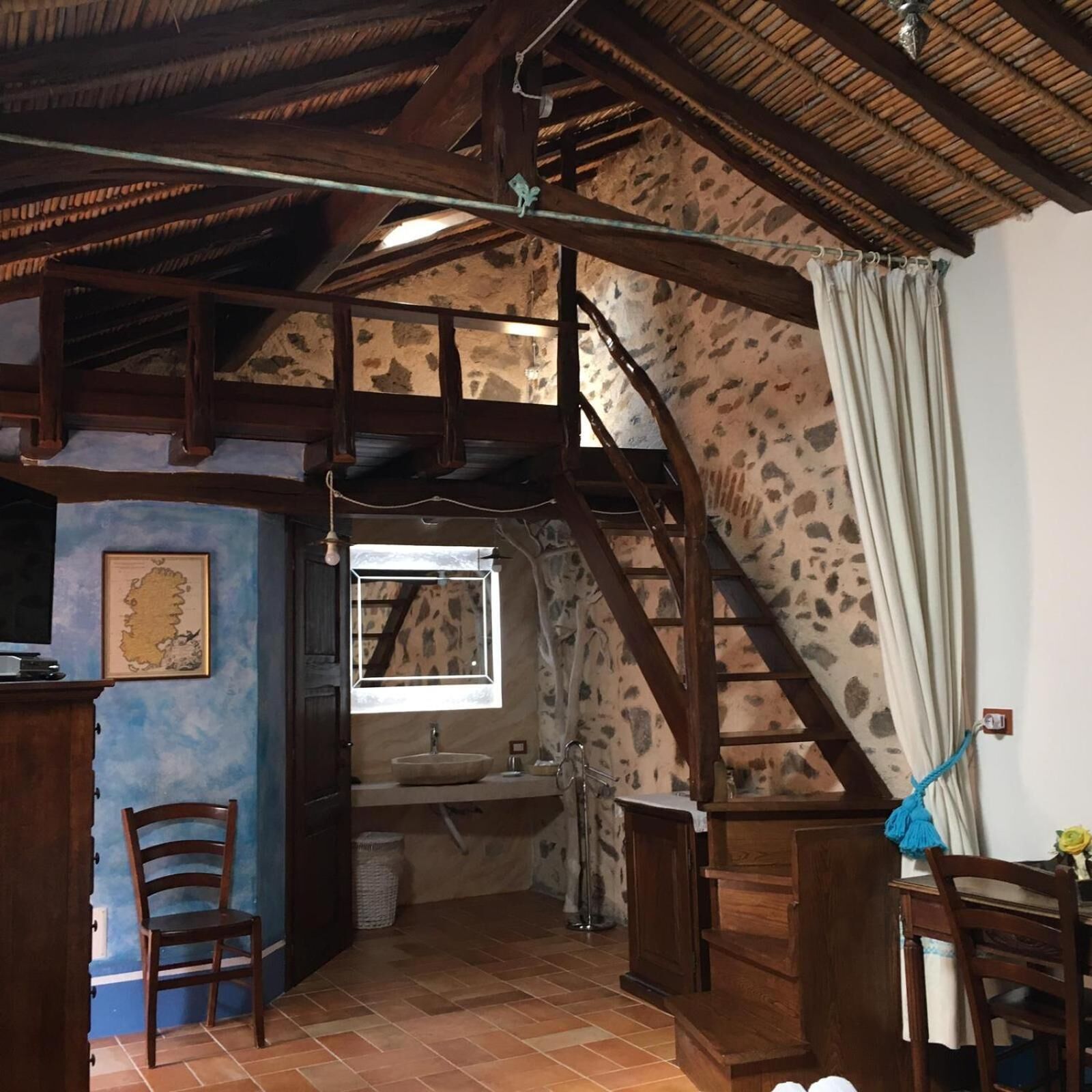 Una habitación rústica con vigas de madera a la vista, pared de piedra, un lavamanos pequeño y una silla.