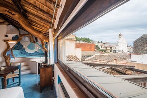Habitación con vista a un paisaje urbano, con piso de baldosas, techo de madera y ventana con vistas a edificios.