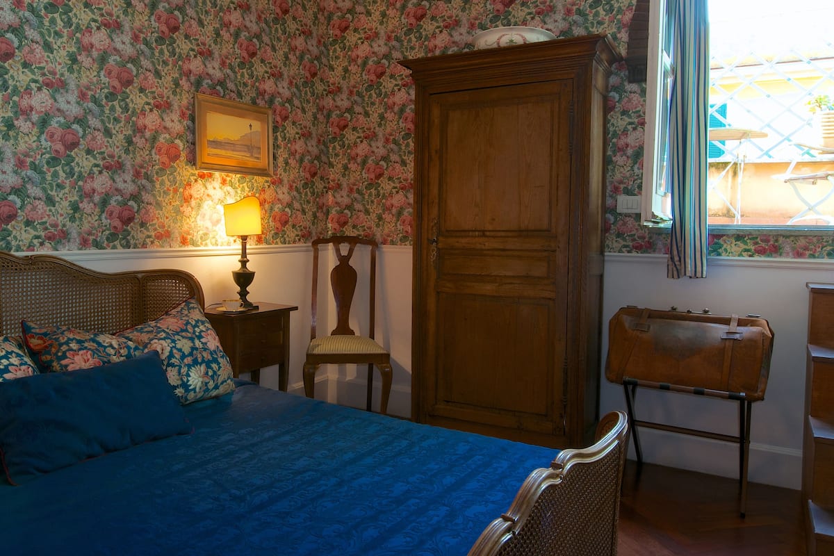 Un dormitori amb paper pintat floral, un armari de fusta, un llit amb roba de llit blava, una nitlínia amb una làmpada, i una finestra amb vista a l'exterior.