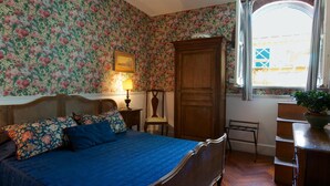 Een slaapkamer met bloemenbehang, een houten garderobe, een bed met blauw beddengoed, een nachtkastje met een lamp en een raam met uitzicht op een gebouw.