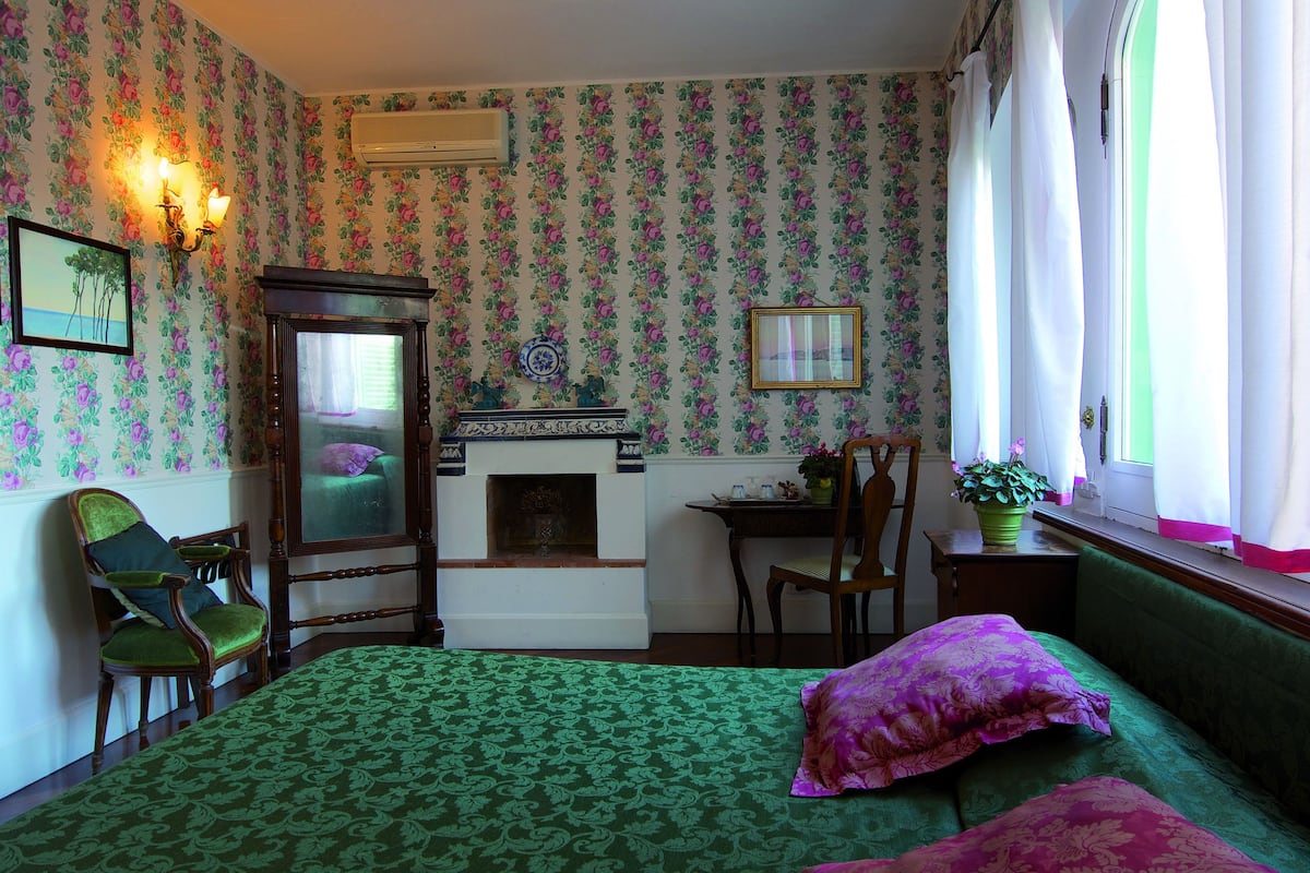 Una habitació amb paper pintat floral, un llit de color verd amb dibuixos, una llar de foc, un escriptori i una cadira.