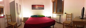 Una habitación pequeña y bien iluminada con una cama, un escritorio, una silla, un radiador y un cuadro de una puesta de sol sobre el océano.