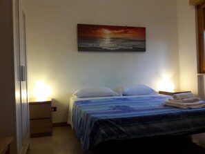 Un dormitorio con cama, dos lámparas de noche, una mesita de noche, un cuadro de una puesta de sol sobre el océano y una ventana con persianas.