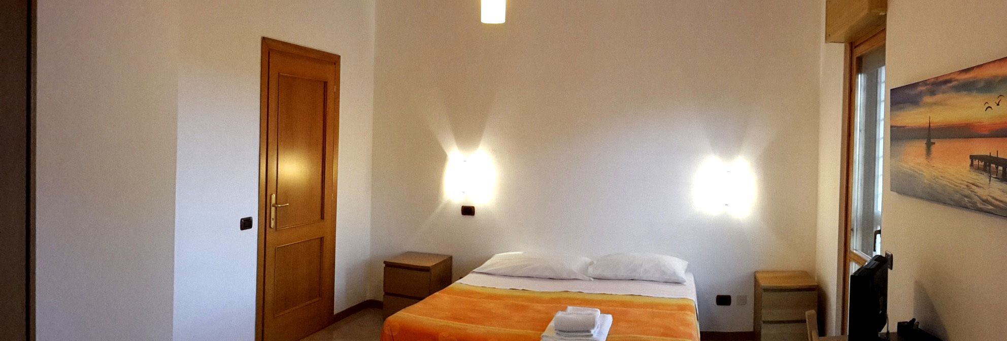 A hotel room with a bed, a door, a nightstand, a wall-mounted light, and a painting on the wall.