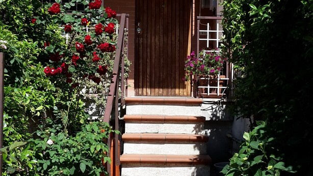 A building entrance with a wooden door, steps leading up to a porch, and a garden with red roses and purple flowers. - Nuova Fiera Aparthouse (Rome)