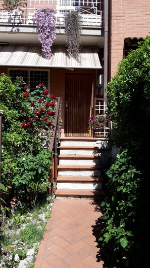 Fachada de un edificio con puerta de madera, escalones que conducen a un porche, y un jardín con rosas rojas y flores moradas.