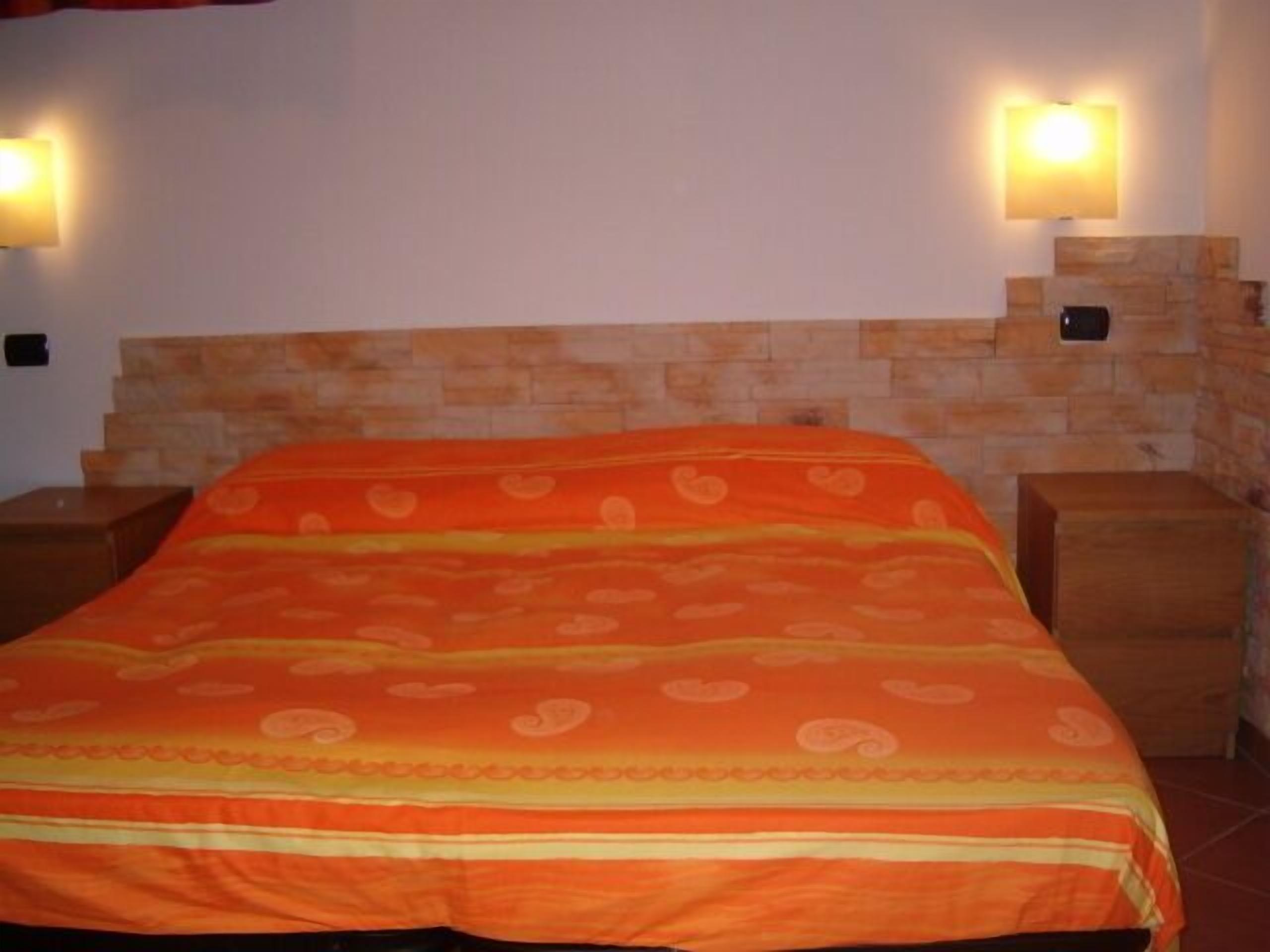Uma cama com colcha laranja estampada, duas luminárias de cabeceira e cabeceira de pedra.